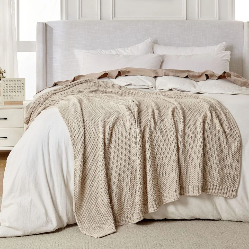 Beige knit throw blanket draped across a white bed in a neutral modern bedroom.
