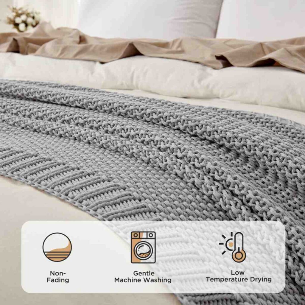 A textured grey Joydeco chunky knit throw blanket draped at the foot of a bed with white linens.