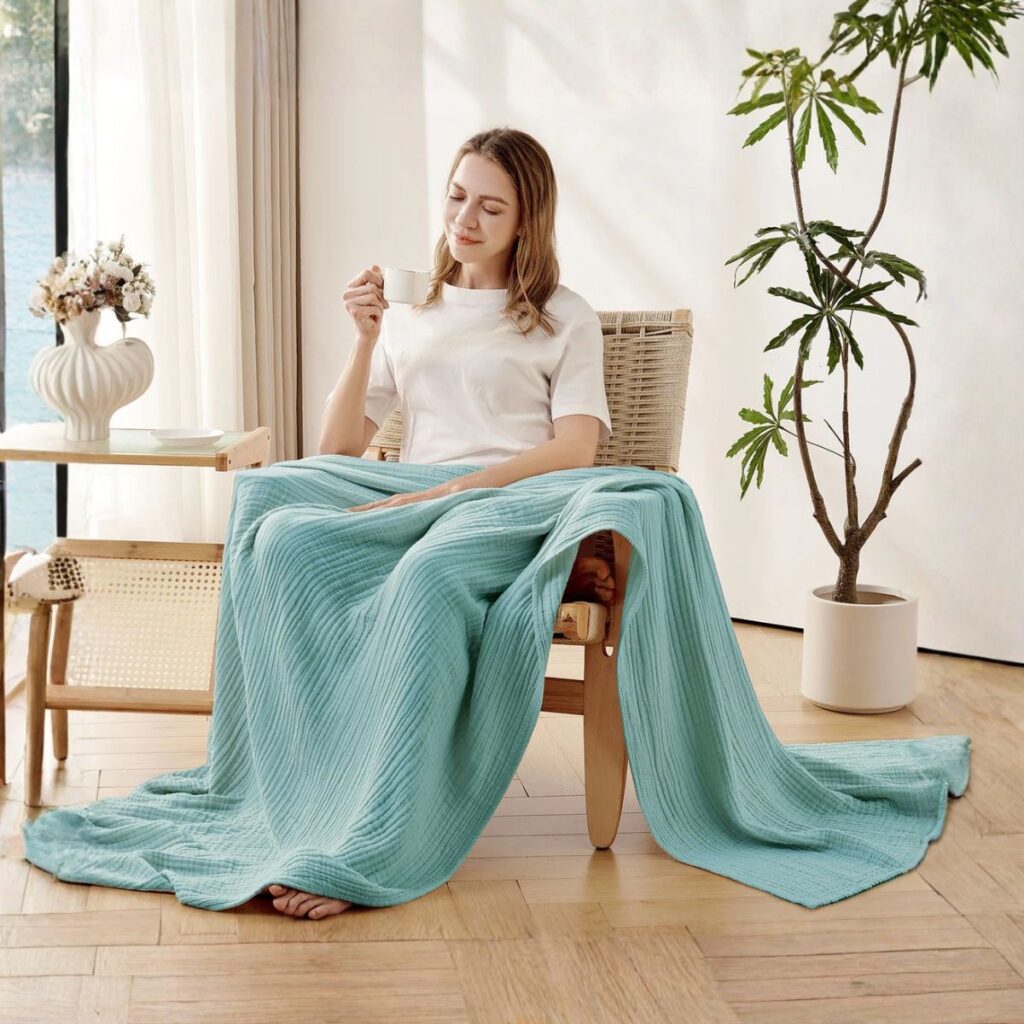 A woman sitting on a chair wrapped in a soft light blue Joydeco throw blanket, highlighting the cozy texture and calming color of the fabric.