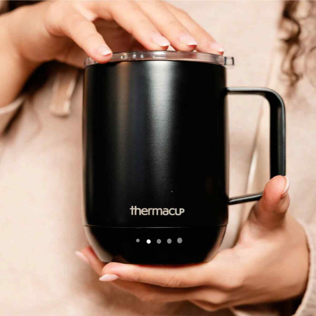 Hands holding a matte black Thermacup smart mug, showcasing the temperature indicator lights and the ergonomic handle design.