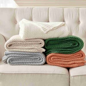 A stack of Joydeco chunky cable knit blankets in white, beige, blue, and orange.